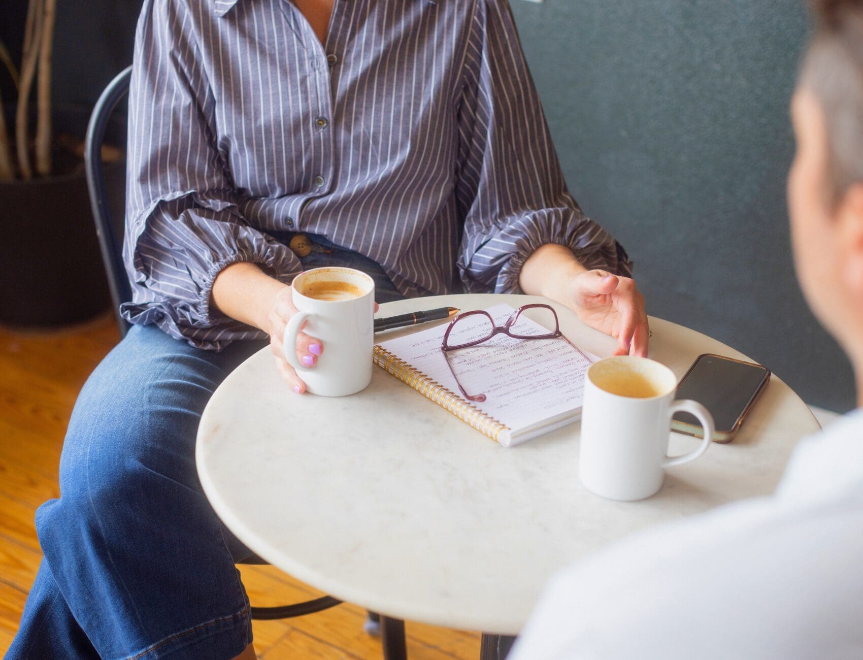 Two people talking over coffee at a table with a notebook, glasses and phone