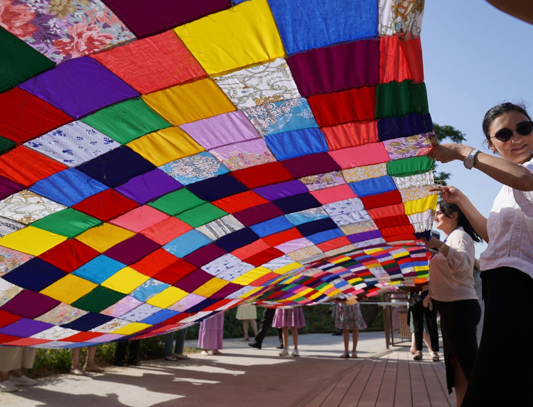 People hold up a large, colorful patchwork quilt outdoors.