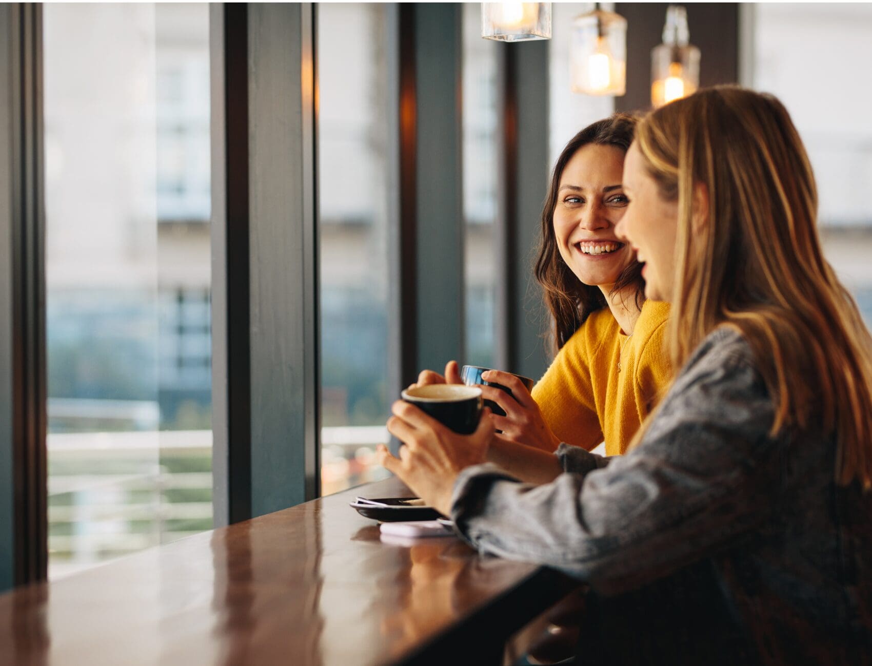 Two women smiling and chatting over coffee at a café counter beside large windows.